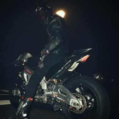A motorcycle rider in a leather jacket sitting on a sports bike at night.